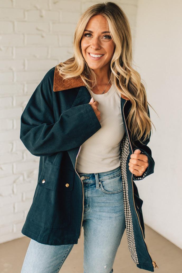 Woman wearing a dark blue coat over a white shirt and blue jeans, standing against a white brick wall.