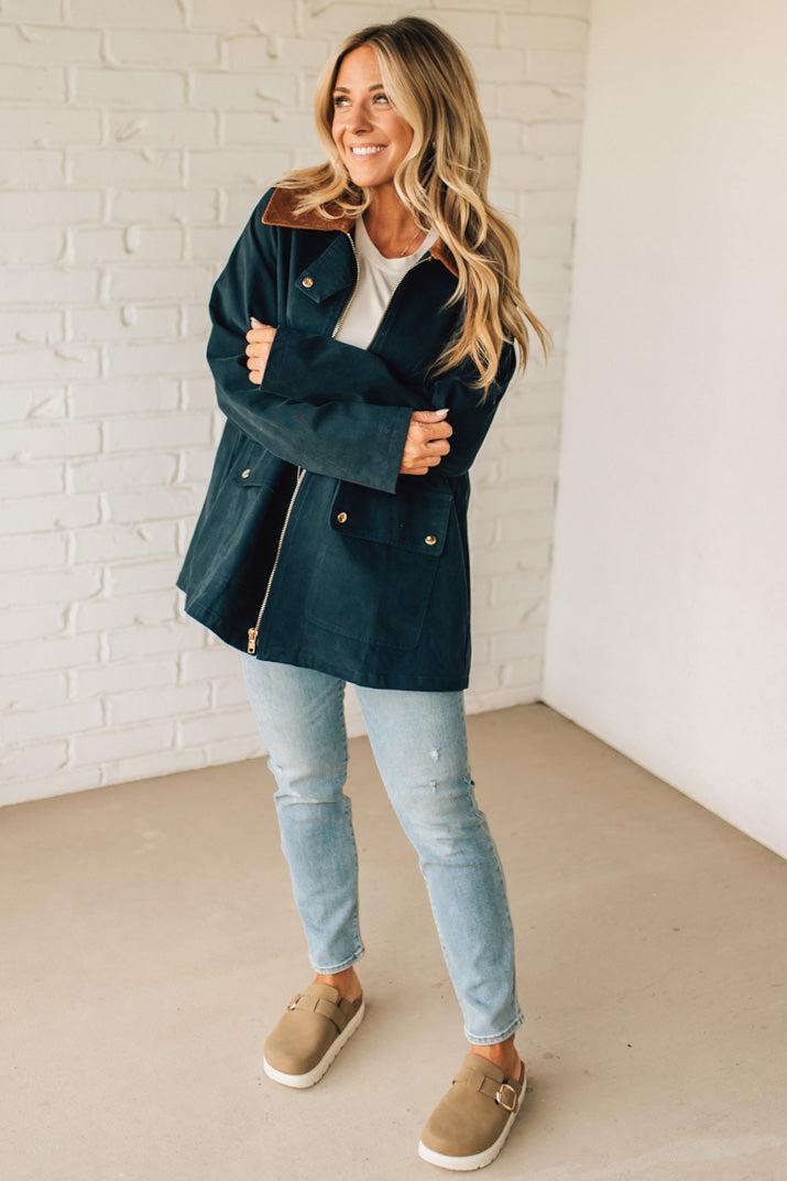 Woman wearing a dark blue coat, light blue jeans, and beige shoes against a white brick wall.