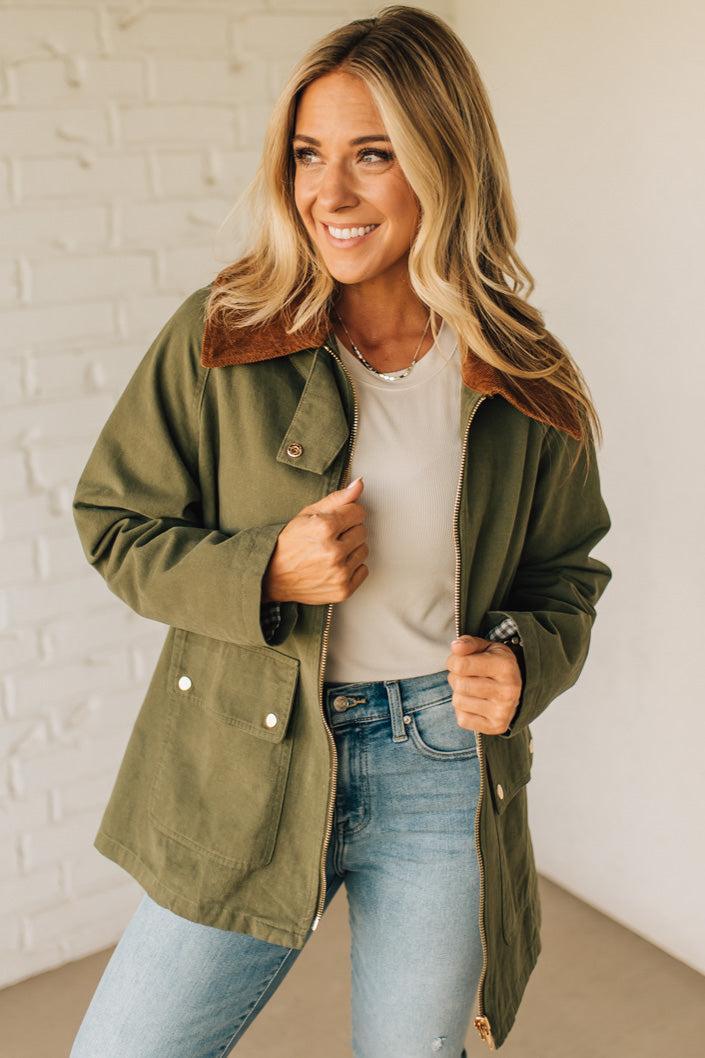 Woman wearing an olive green jacket with a brown collar, white shirt, and blue jeans against a white brick wall.
