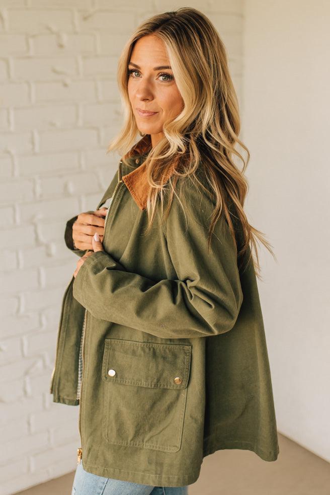 Woman wearing an olive green jacket against a white brick wall.