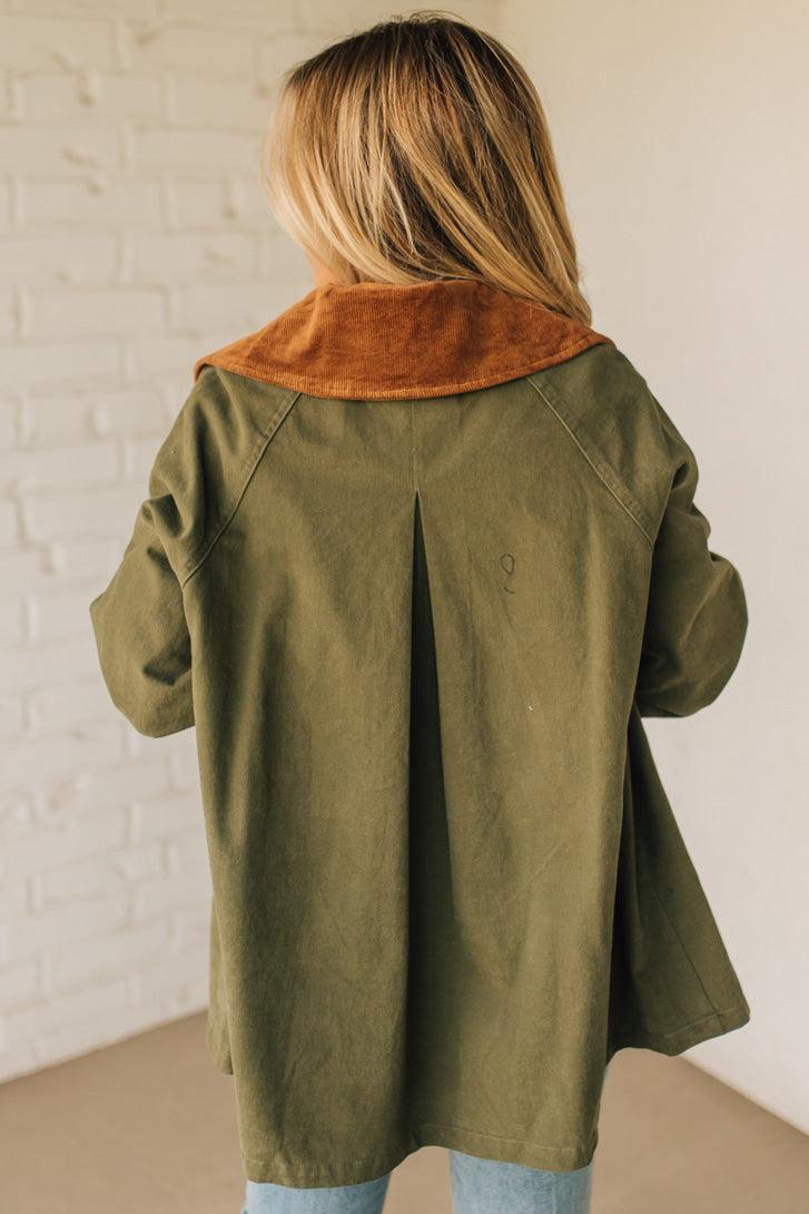 Person wearing an olive green coat with a brown collar against a white brick wall.