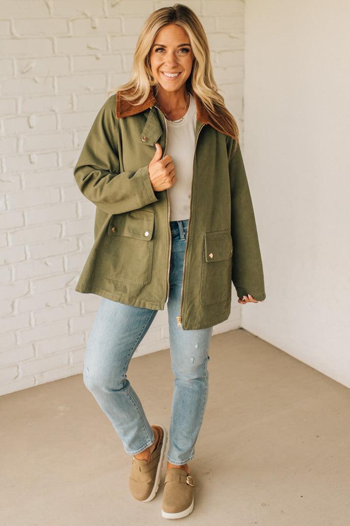 Woman wearing an olive green jacket, white shirt, blue jeans, and brown shoes against a white brick wall.