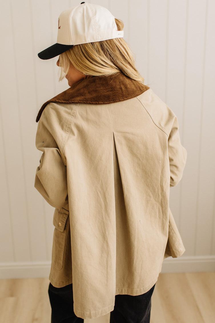 Person wearing a beige trench coat with a brown collar and a white cap, standing against a light-colored wall.