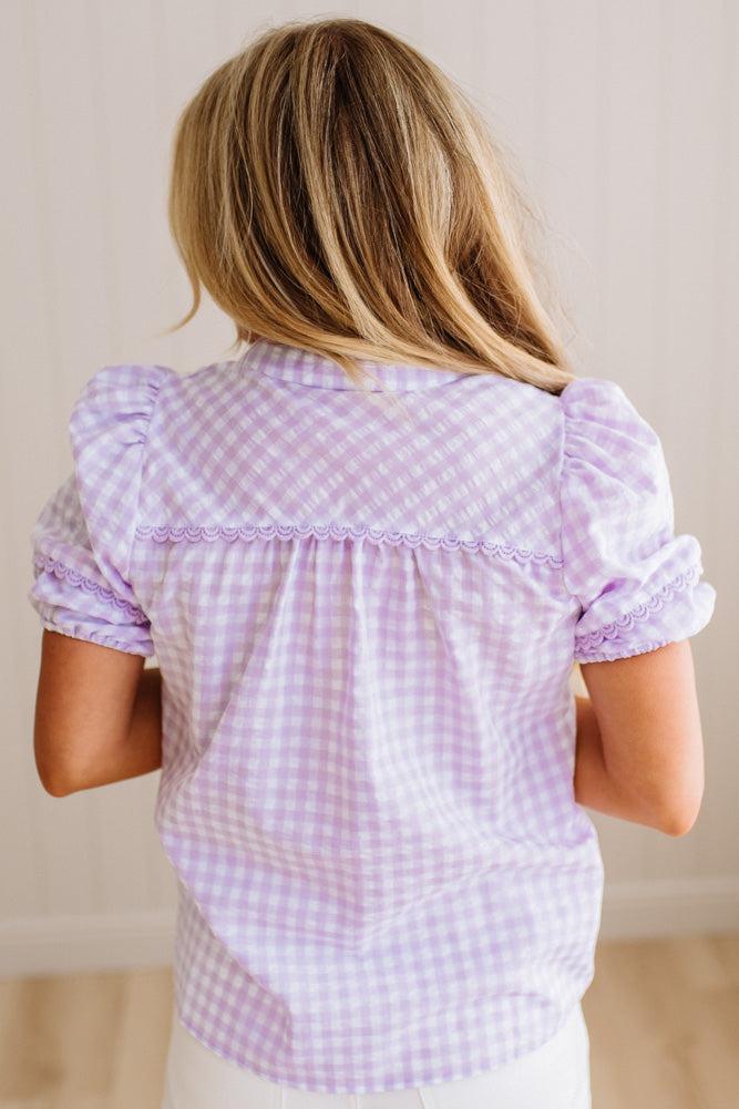Blonde woman wearing a lavender and white gingham top with scallop edge trim and puff sleeves.