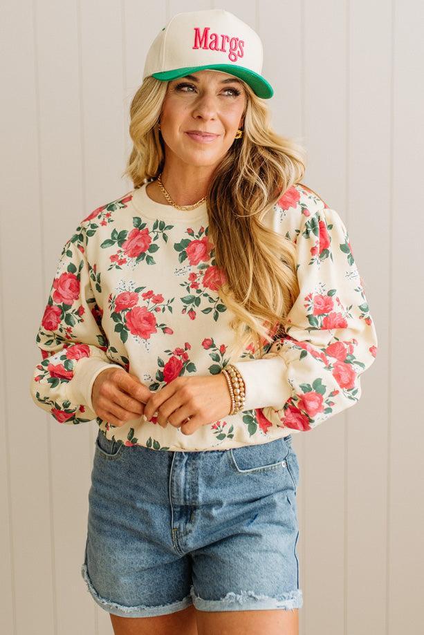 Woman wearing a floral sweatshirt, denim shorts, and a cap with 'Margs' text.