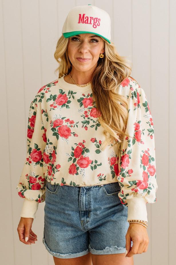 Woman wearing a floral sweatshirt, denim shorts, and a cap with 'Margs' text.