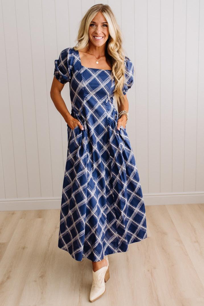 A woman modeling a blue and white patterned midi dress with puff sleeves, a square neckline, and side pockets, standing in front of a white paneled wall and light wood floor.