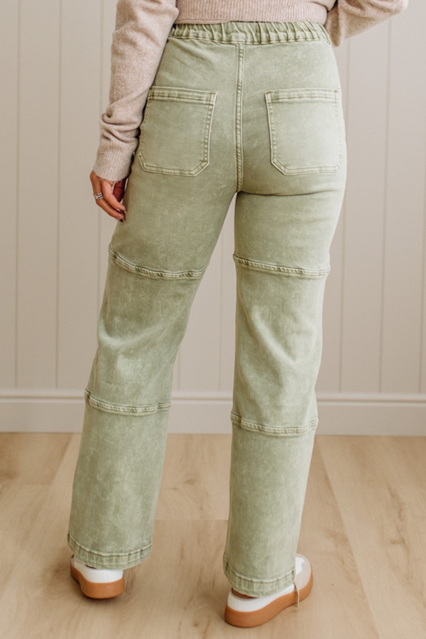 Person wearing light green jeans standing on a wooden floor with a white paneled wall in the background.