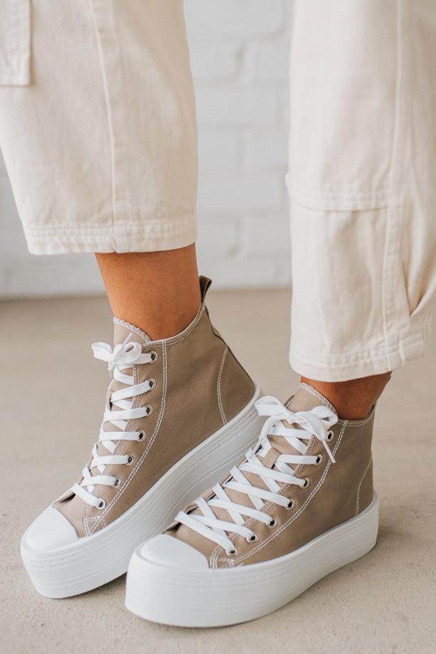 Beige high-top sneakers with white laces and soles worn with light-colored pants on a neutral background.