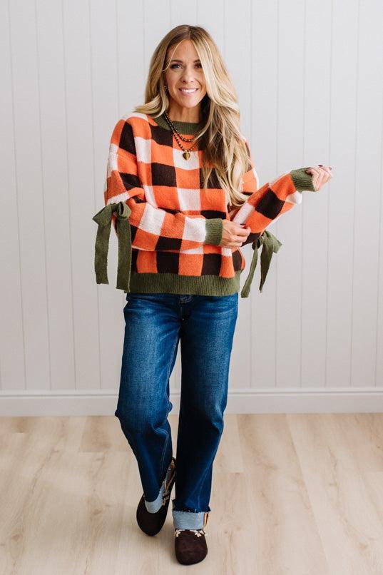 Blonde woman wearing a orange, brown and cream check sweater with olive green ribbed trim and bows at the elbow.