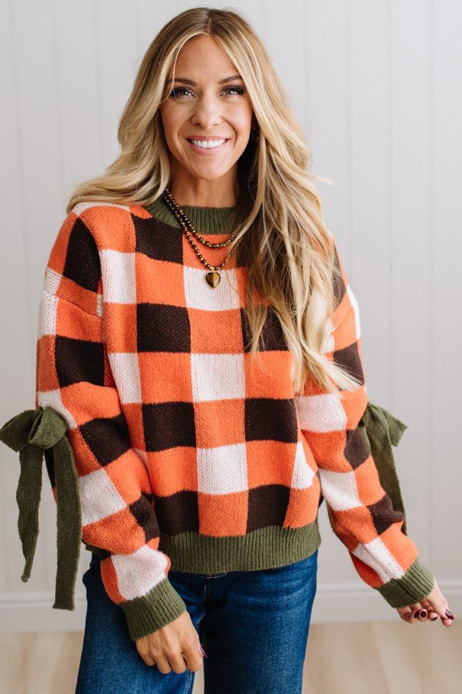 Blonde woman wearing a orange, brown and cream check sweater with olive green ribbed trim and bows at the elbow.
