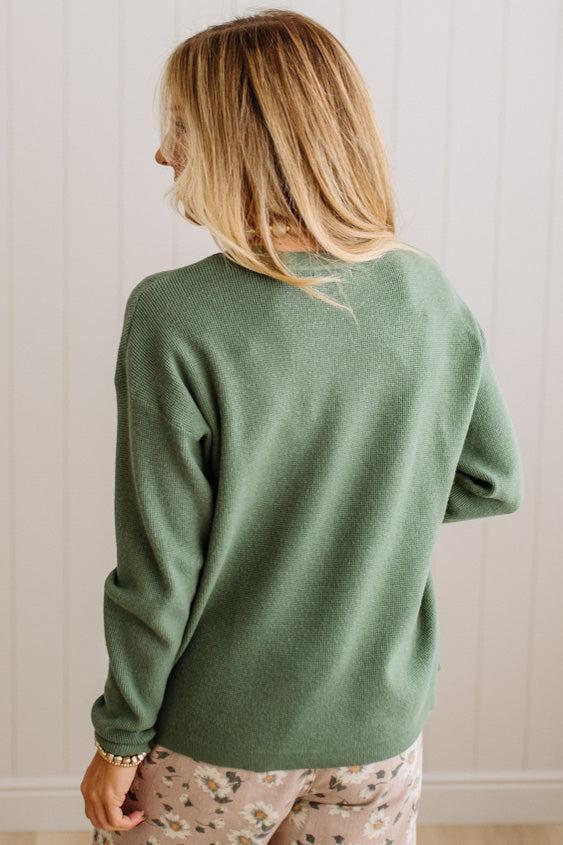 A woman with blonde hair is shown from behind wearing a sage green knit sweater and light pink floral pajama pants against a white paneled wall.