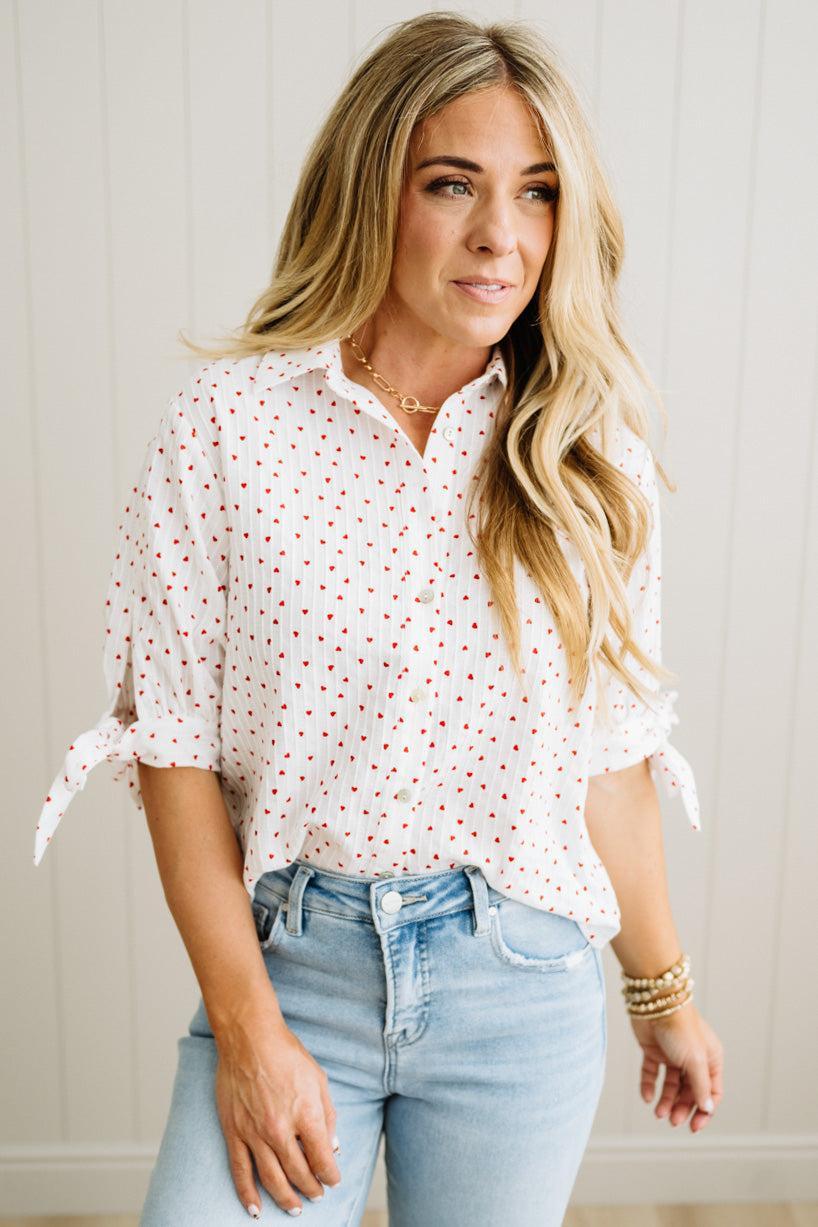 Woman wearing a white blouse with red heart patterns and light blue jeans against a neutral background