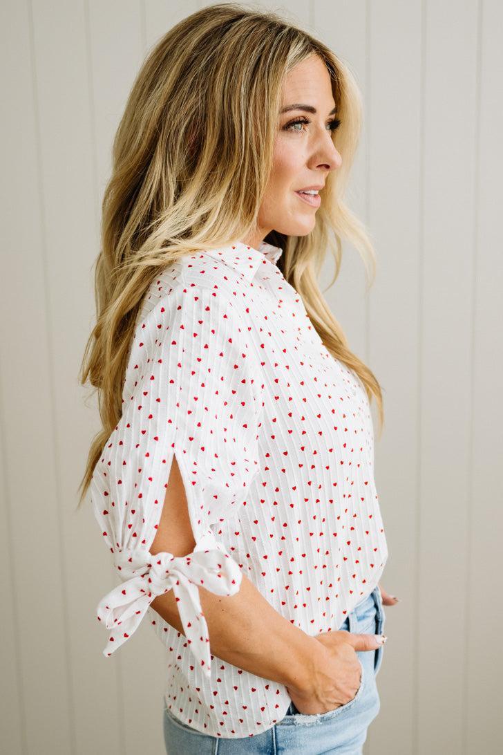 Woman wearing a white blouse with red mini heart patterns and light blue jeans against a neutral background