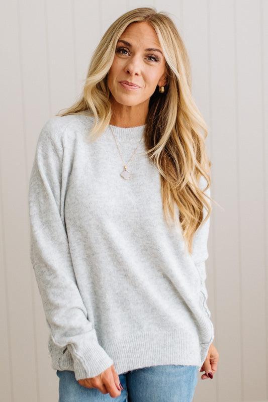 BLONDE WOMAN WEARING A HIGH-LOW HEM, LONG SLEEVE, SUPER SOFT, OVERSIZED CREW NECK SWEATER WITH RIB DETAILS AND STANDING IN A STUDIO WITH LIGHT WOOD FLOOR AND PANELED WALL BEHIND.