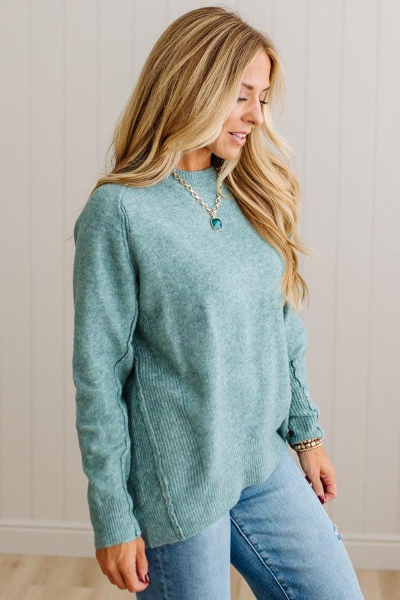 BLONDE WOMAN WEARING A HIGH-LOW HEM, LONG SLEEVE, SUPER SOFT, OVERSIZED CREW NECK SWEATER WITH RIB DETAILS AND STANDING IN A STUDIO WITH LIGHT WOOD FLOOR AND PANELED WALL BEHIND.