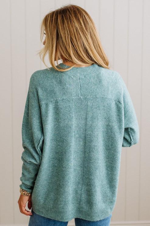 BLONDE WOMAN WEARING A HIGH-LOW HEM, LONG SLEEVE, SUPER SOFT, OVERSIZED CREW NECK SWEATER WITH RIB DETAILS AND STANDING IN A STUDIO WITH LIGHT WOOD FLOOR AND PANELED WALL BEHIND.