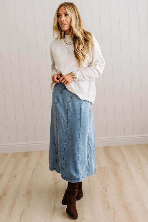 BLONDE WOMAN WEARING A HIGH-LOW HEM, LONG SLEEVE, SUPER SOFT, OVERSIZED CREW NECK SWEATER WITH RIB DETAILS AND STANDING IN A STUDIO WITH LIGHT WOOD FLOOR AND PANELED WALL BEHIND.