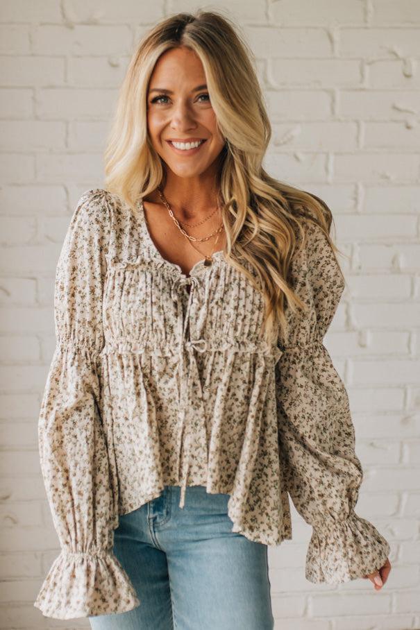 Woman wearing a patterned blouse with long sleeves against a white brick wall.