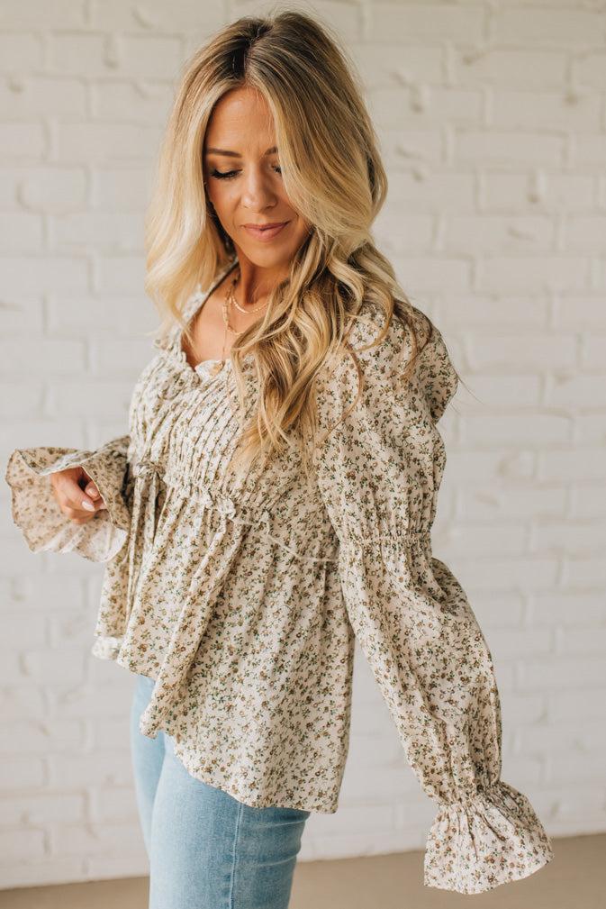 Woman wearing a floral blouse with long sleeves against a white brick wall.