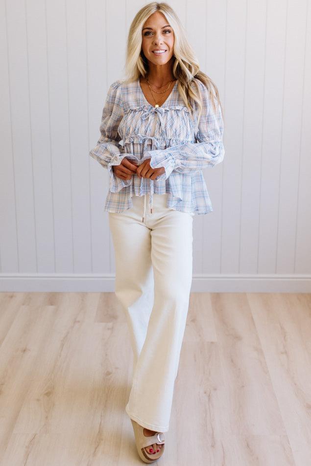 A woman modeling a light blue and white plaid long-sleeve blouse with a ruffled tie neckline and ruffled cuffs, paired with cream-colored wide-leg pants, standing in front of a white paneled wall and light wood floor.