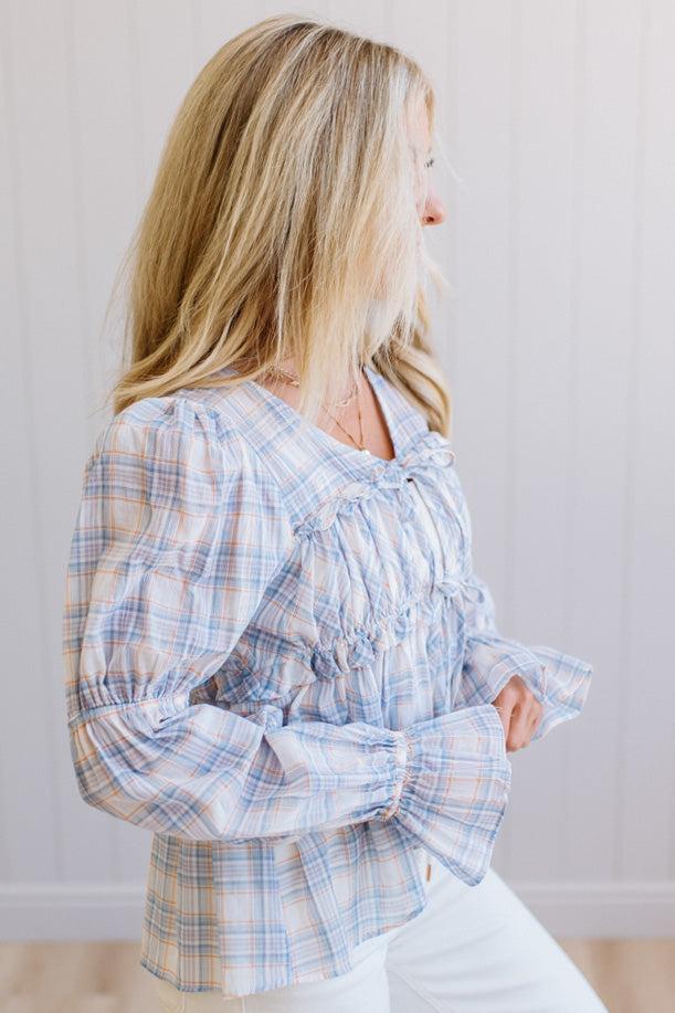 A woman wearing a light blue and white plaid long-sleeve blouse with ruffles, shown in profile against a light paneled wall.