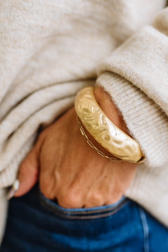 Woman in striped top and light jeans with her hand an a pocket and focused on a large matte gold bangle.