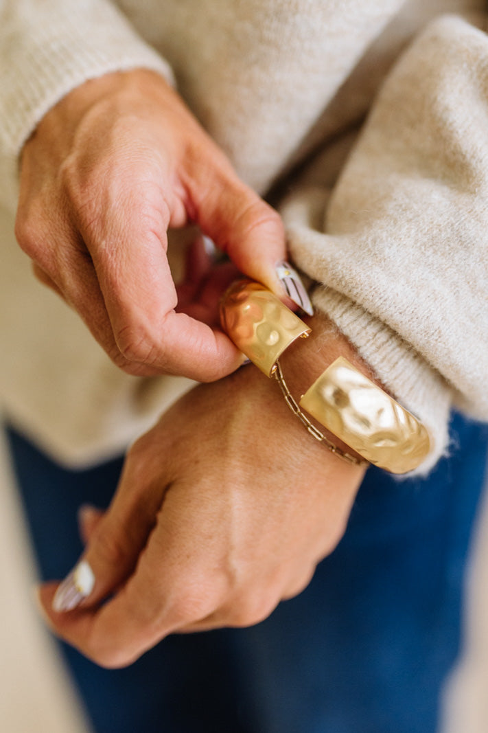 Woman in striped top and light jeans with her hand an a pocket and focused on a large matte gold bangle.