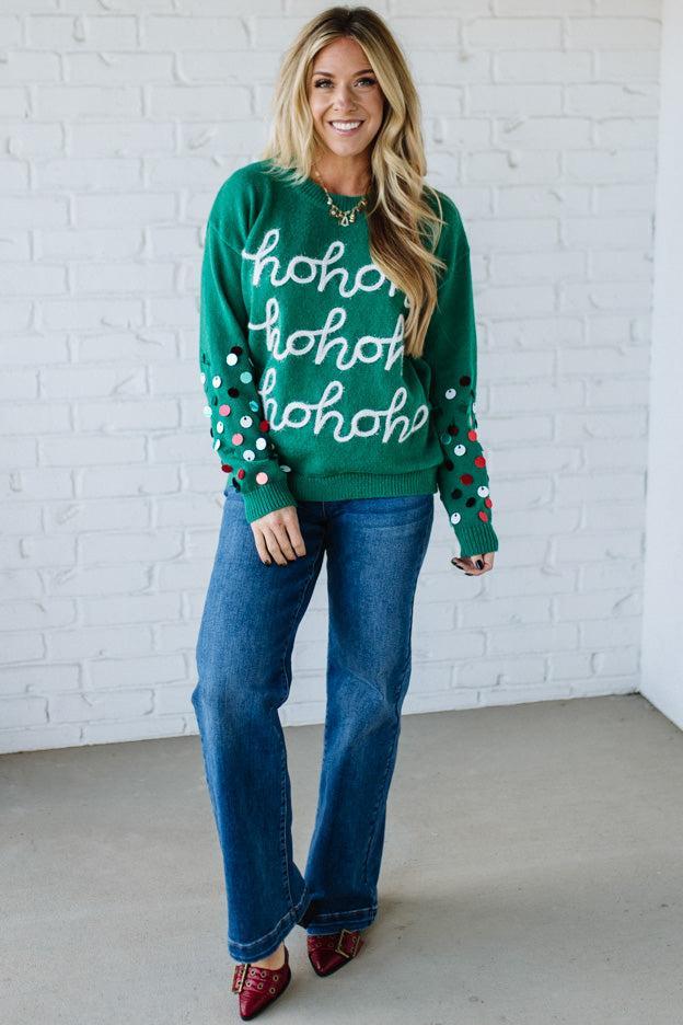 Woman wearing a green sweater with 'ho ho ho' text and blue jeans against a white brick wall.