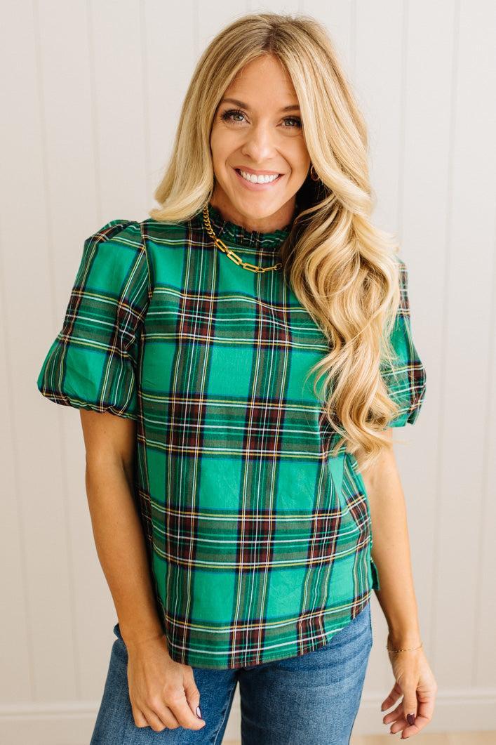 Woman wearing a green plaid top with ruffled sleeves against a white wall.