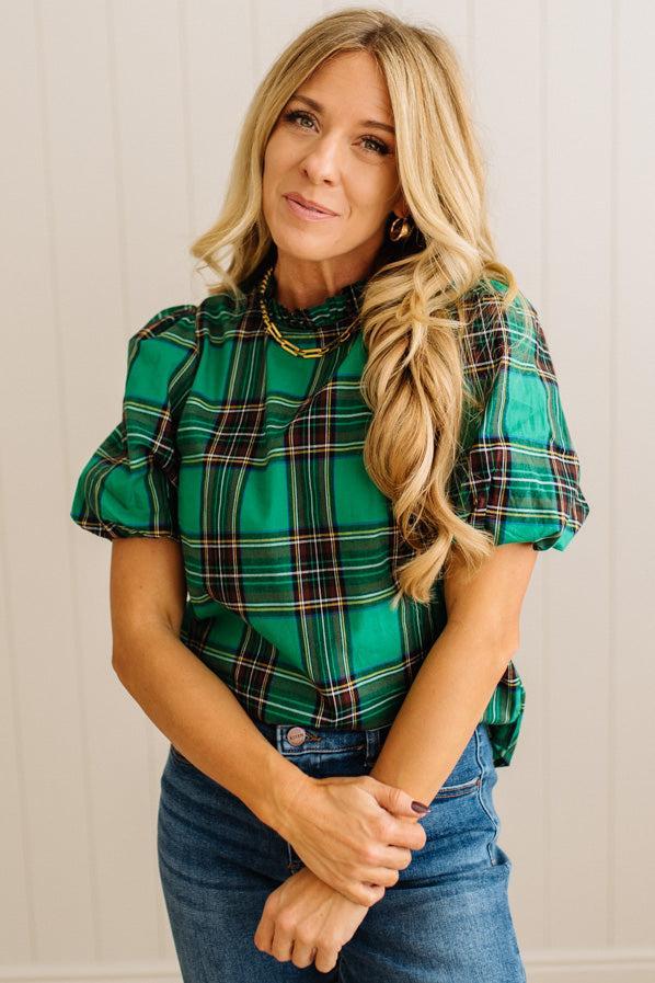 Woman wearing a green plaid shirt and blue jeans against a white wall.