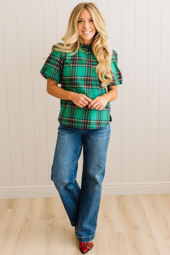 Woman wearing a green plaid top and blue jeans standing against a white paneled wall.