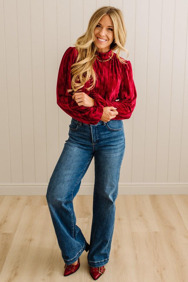 Woman wearing a red velvet blouse and blue jeans standing against a beige wall.