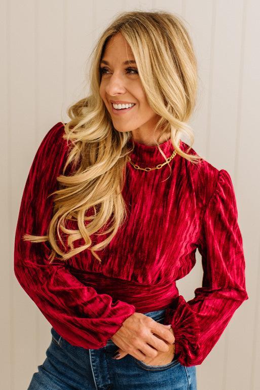 Woman wearing a red velvet blouse with long sleeves and blue jeans against a neutral background