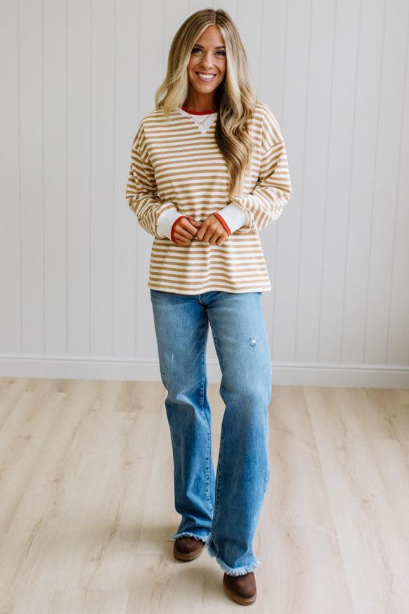 BLONDE WOMAN WEARING A CAMEL AND WHITE STRIPED CREW NECK PULLOVER WITH WHITE AND DARK ORANGE KNIT RIB BAND TRIMMED, IT IS LONG SLEEVED WITH A RELAXED FIT. SHE IS STANDING ON A LIGHT WOOD FLOOR WITH LIGHT BACKGROUND.