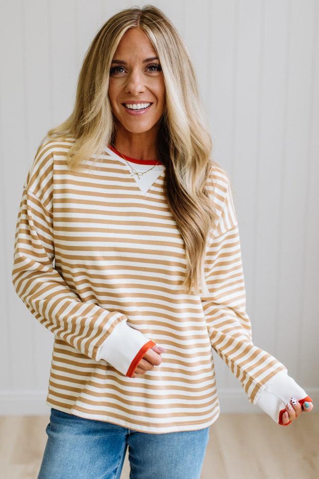 BLONDE WOMAN WEARING A CAMEL AND WHITE STRIPED CREW NECK PULLOVER WITH WHITE AND DARK ORANGE KNIT RIB BAND TRIMMED, IT IS LONG SLEEVED WITH A RELAXED FIT. SHE IS STANDING ON A LIGHT WOOD FLOOR WITH LIGHT BACKGROUND.