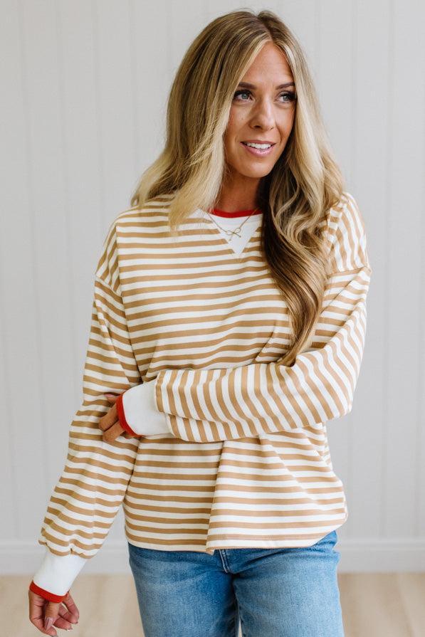 BLONDE WOMAN WEARING A CAMEL AND WHITE STRIPED CREW NECK PULLOVER WITH WHITE AND DARK ORANGE KNIT RIB BAND TRIMMED, IT IS LONG SLEEVED WITH A RELAXED FIT. SHE IS STANDING ON A LIGHT WOOD FLOOR WITH LIGHT BACKGROUND.