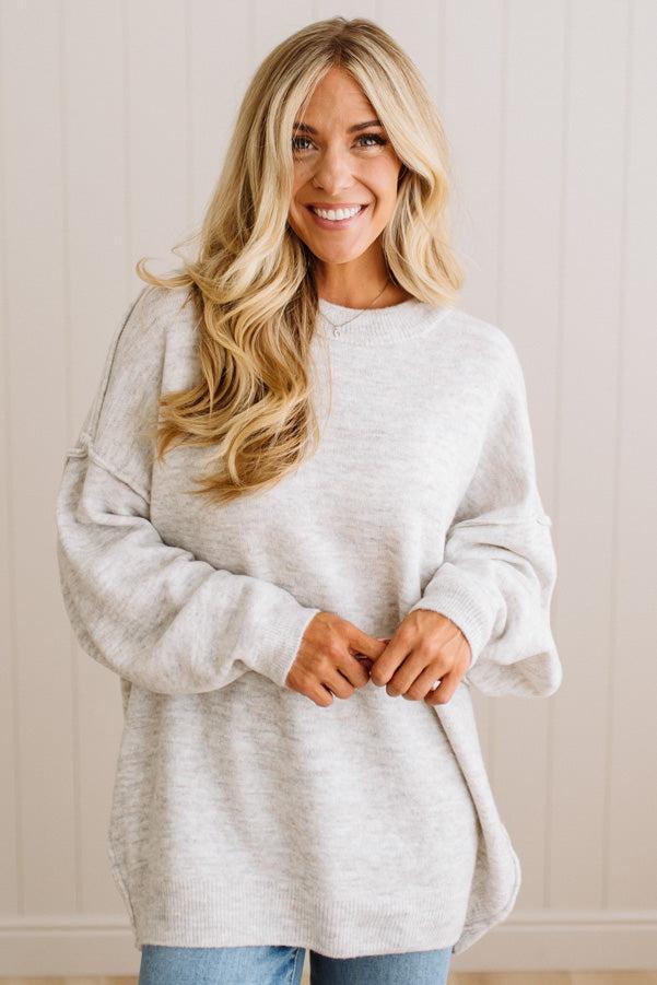 A smiling blonde woman wearing a light heather gray oversized crewneck sweatshirt and blue jeans, standing against a light beige paneled wall.