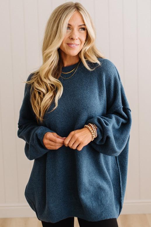 A woman with long blonde hair wearing an oversized, dusty slate-blue knit sweater, standing against a white paneled wall.