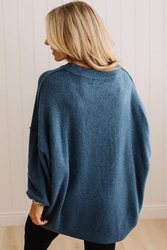 A woman with blonde hair is shown from behind wearing a loose-fitting, oversized blue knit sweater. She is standing in front of a white paneled wall and light wood floor.