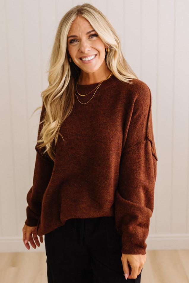 A smiling blonde woman wearing a fuzzy brown long-sleeve sweater and black pants, standing against a light beige paneled wall.
