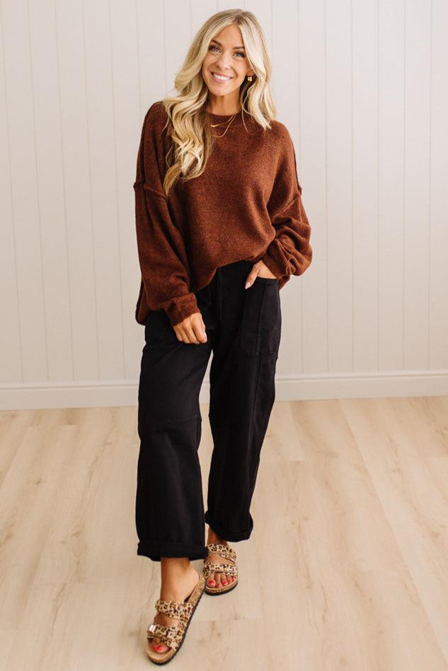A woman modeling a brown crewneck knit sweater and black wide-leg pants, standing on a light wood floor against a white paneled wall.