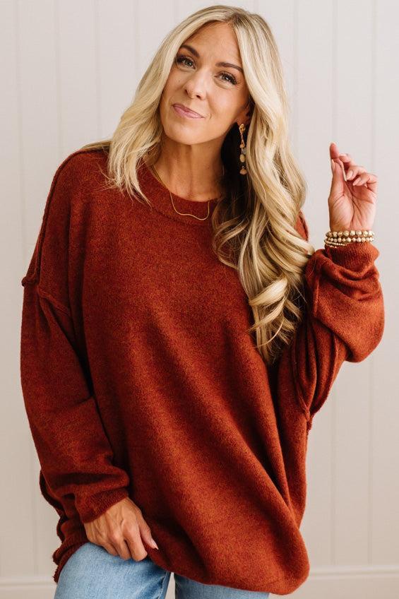 A smiling blonde woman wearing a rust oversized crewneck sweatshirt and blue jeans, standing against a light beige paneled wall.