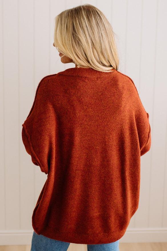 A woman viewed from behind wearing a loose, oversized rust-orange knit sweater and blue jeans, standing against a white paneled wall.