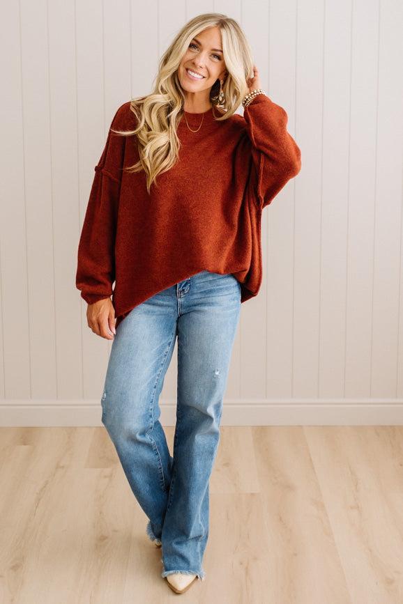 A woman wearing a rust-colored knit sweater and light-wash blue jeans, standing in a room with light wood floors and white paneled walls.