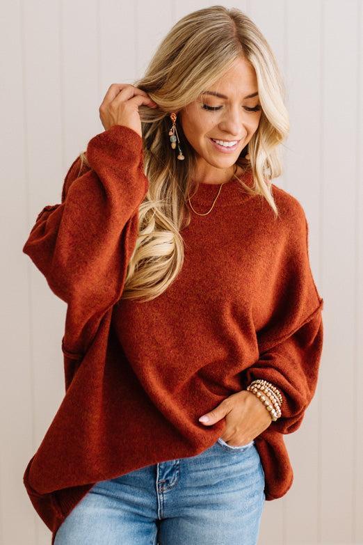 A woman wearing a cozy, oversized rust-orange knit sweater and light-wash blue jeans, standing against a light beige paneled wall.