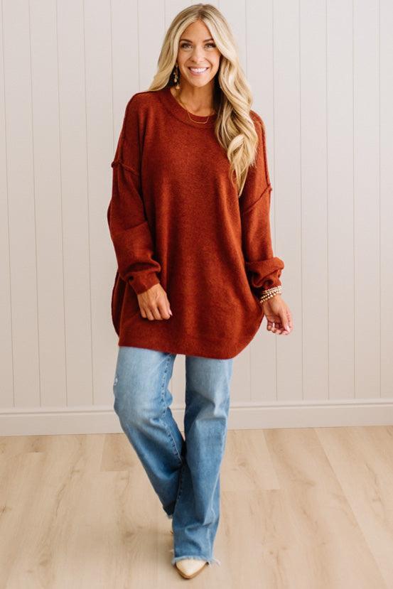 A smiling blonde woman wearing a rusty oversized crewneck sweatshirt and blue jeans, standing against a light beige paneled wall.