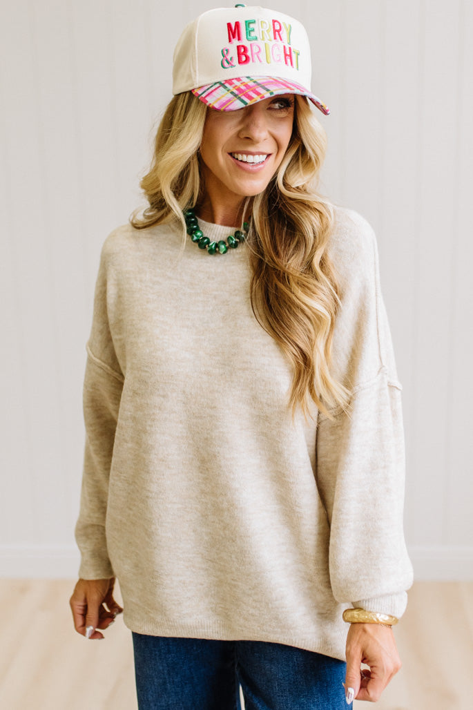 A woman with long blonde hair wearing an oversized light heather beige crewneck sweatshirt and blue jeans, standing against a light beige paneled wall.