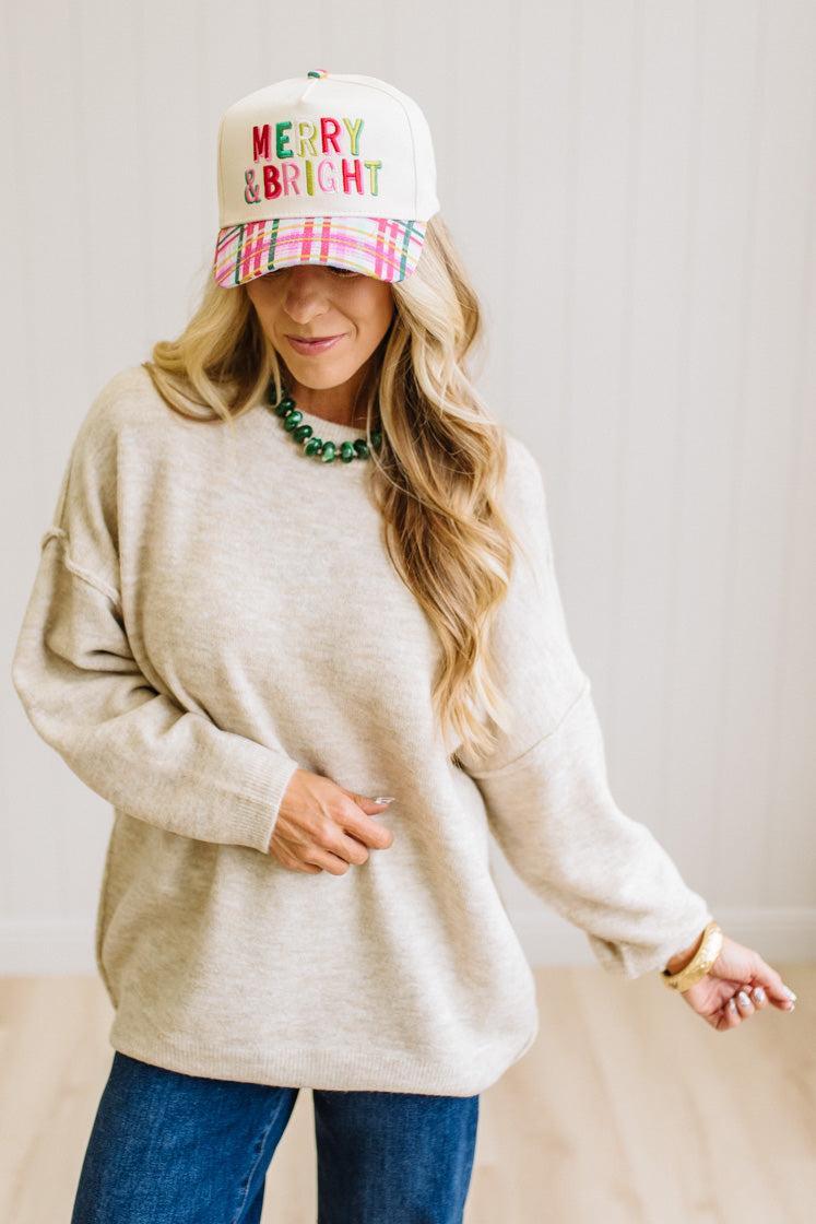 A woman with long blonde hair wearing an oversized light heather beige crewneck sweatshirt and blue jeans, standing against a light beige paneled wall.