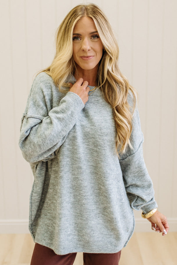 A woman with long blonde hair wearing an oversized light heather gray crewneck sweatshirt and blue jeans, standing against a light beige paneled wall.